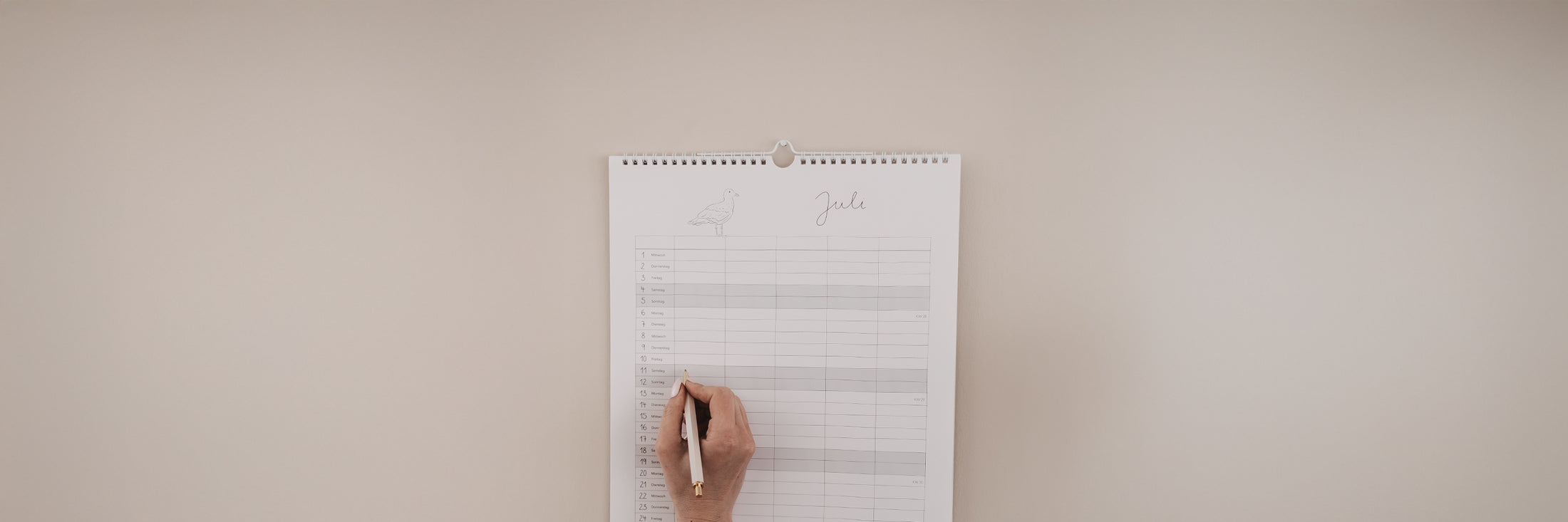 Familienkalender 2026 mit liebevoll gestalteten Illustrationen hängt an der Wand, jemand trägt Termine mit unserem cremefarbenen Kugelschreiber ein. Oben im Bild steht: „Kalender – für all die schönen Momente“ und unten als Schaltfläche „Zu den Kalendern“