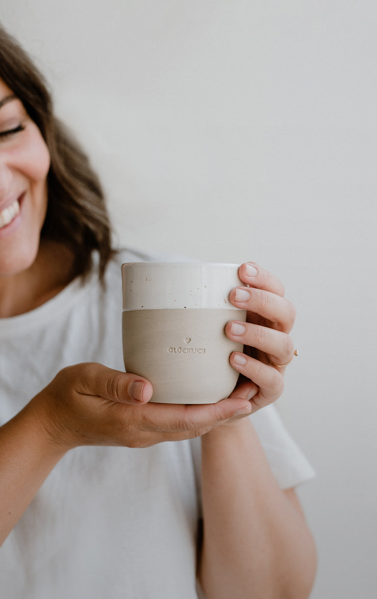 Lächelnde Frau in weißem T-Shirt hält einen hellen Steingutbecher mit Blindprägung ‚Glücklich‘ in beiden Händen vor sich.