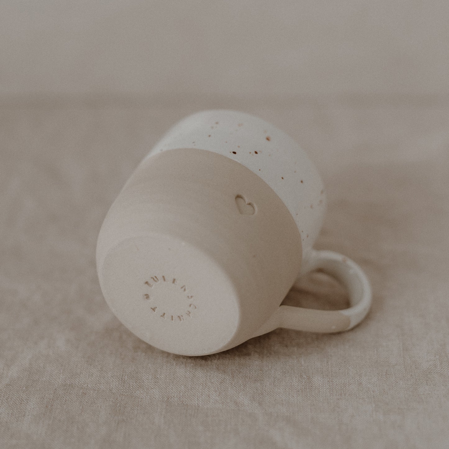 Eine beige-weiße Keramiktasse liegt auf der Seite auf einem Leinentuch. Die obere Hälfte ist glasiert und weiß mit kleinen braunen Sprenkeln, die untere Hälfte matt beige. Ein kleines, eingraviertes Herz ist an der Seite zu sehen. Auf dem Tassenboden steht in einem Kreis: ‚Zuhause bei mir‘.