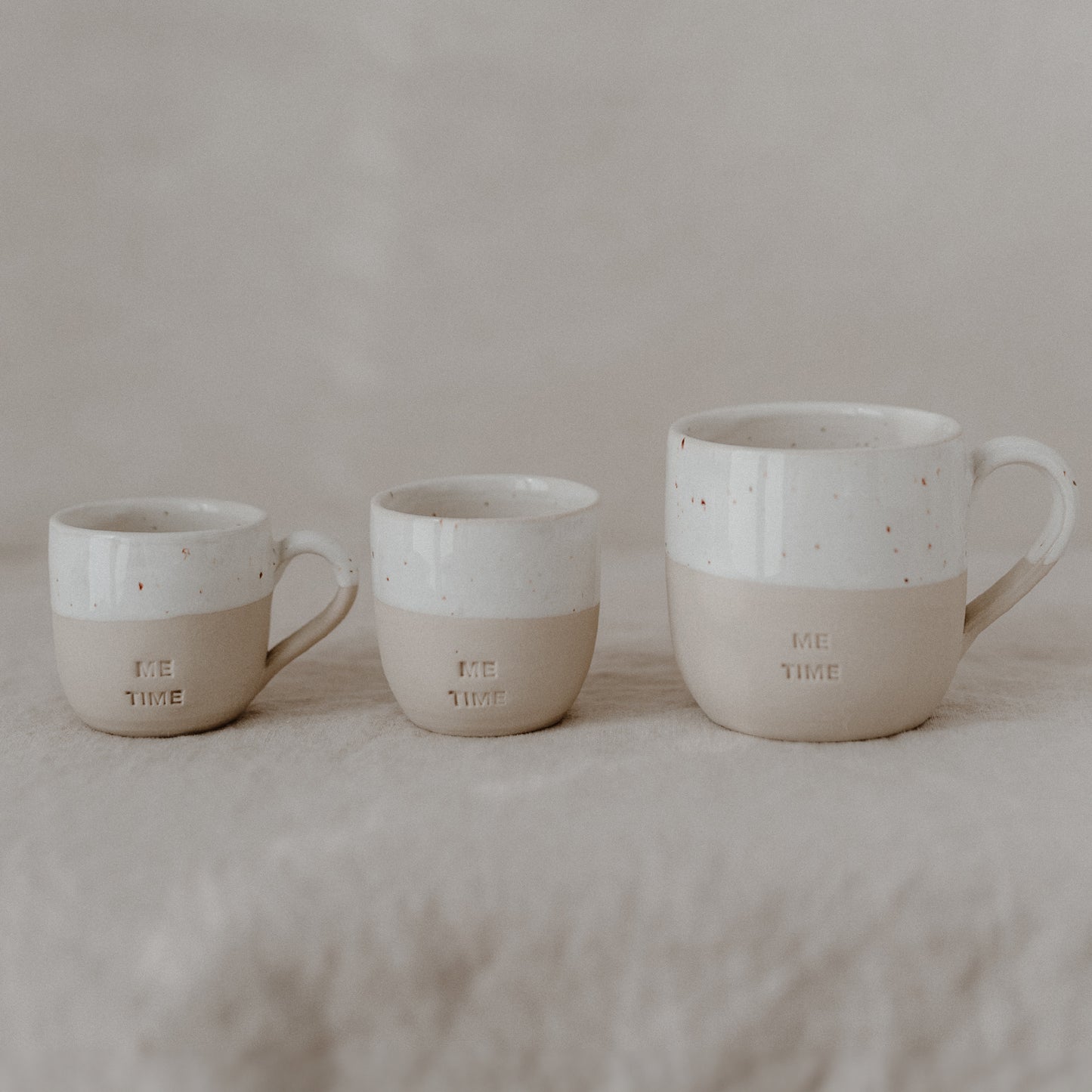 Drei Keramiktassen in unterschiedlichen Größen stehen nebeneinander auf einer beigefarbenen, weichen Stoffoberfläche. Jede Tasse ist in Beige und Weiß gehalten, wobei der obere Teil weiß glasiert ist und kleine braune Sprenkel zeigt. Auf dem unteren, matten Teil ist bei allen Tassen das Wort ‚ME TIME‘ eingeprägt.
