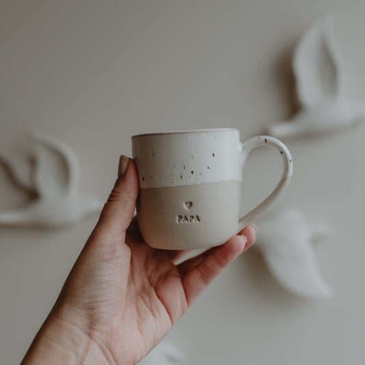 Hand hält eine Cappuccinotasse aus Keramik mit Herzprägung und dem Schriftzug PAPA, vor einem hellen Hintergrund mit Vogelreliefs.