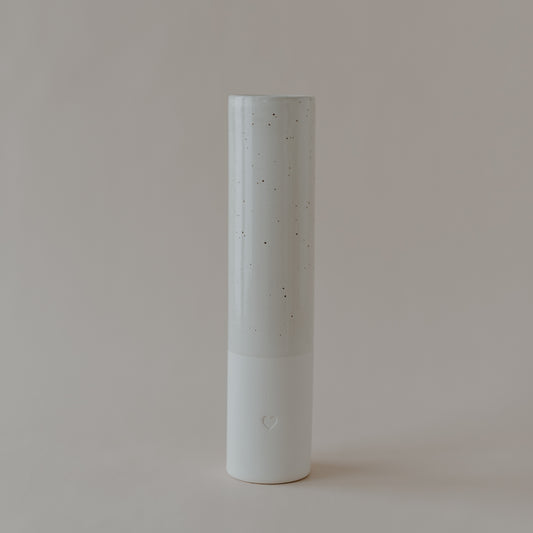 Hohe zylindrische Keramikvase mit gesprenkelter glänzender Oberfläche im oberen Bereich und matter weißer Basis mit eingeprägtem Herzsymbol, stehend vor neutralem Hintergrund.