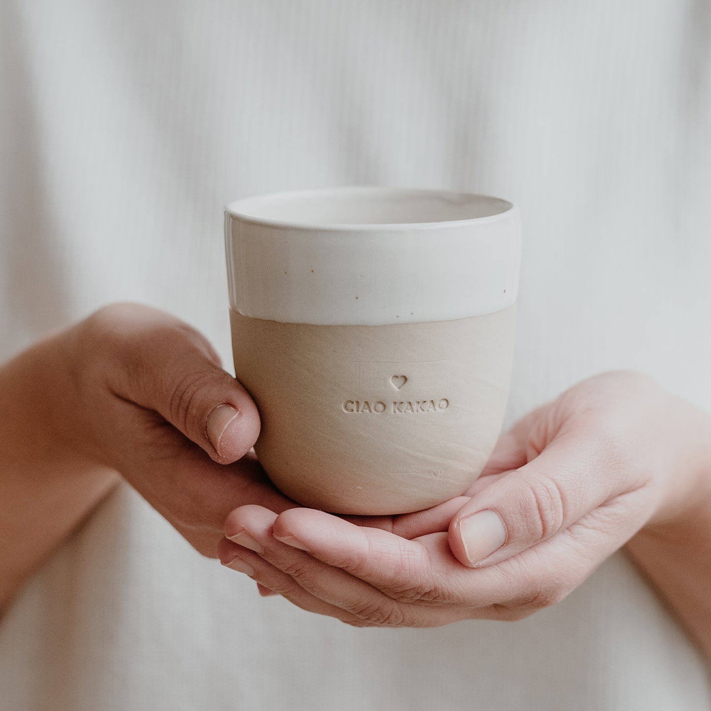Zwei Hände präsentieren einen schlichten Becher mit CIAO KAKAO Prägung vor einem hellen, textilen Hintergrund.