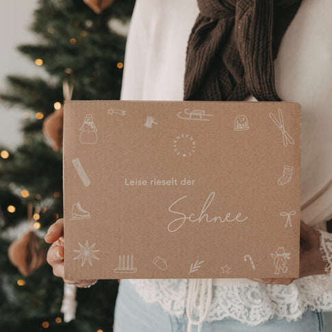Limitierte Winter-Überraschungsbox 2026 von Eulenschnitt mit winterlichen Illustrationen und Schriftzug „Leise rieselt der Schnee“, vor einem geschmückten Weihnachtsbaum gehalten.