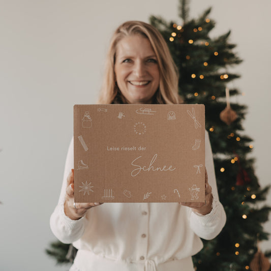 Lächelnde Frau hält die limitierte Winter-Überraschungsbox 2026 von Eulenschnitt mit liebevollen Wintermotiven vor einem beleuchteten Weihnachtsbaum.