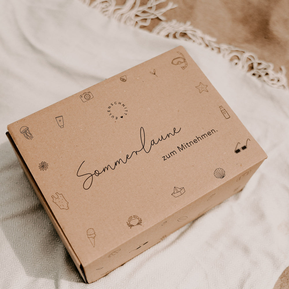 Eine Kraftpapierbox mit der Aufschrift "Sommerlaune zum Mitnehmen" und verschiedenen sommerlichen Details. Im Hintergrund Strand und ein Handtuch.