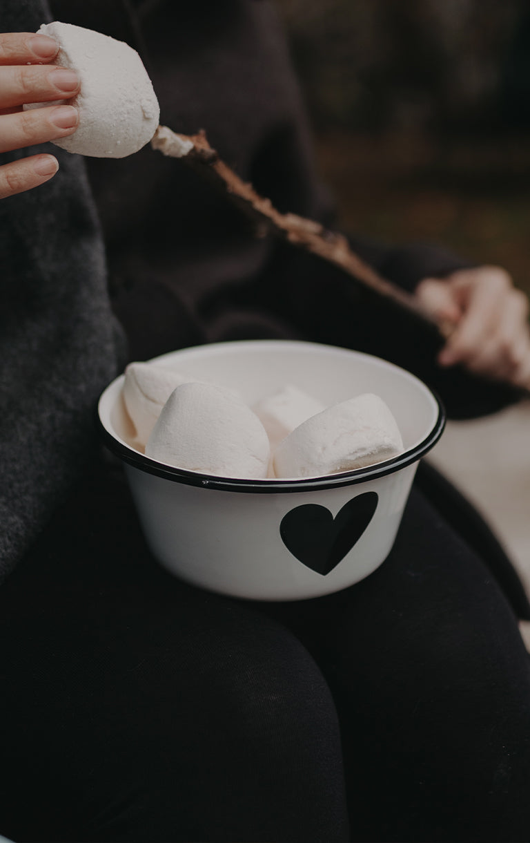 Emaille-Schüssel mit schwarzem Herzmotiv, gefüllt mit Marshmallows, wird auf dem Schoß gehalten. Text im Bild: „Für jedes Abenteuer – Emaille-Geschirr.“