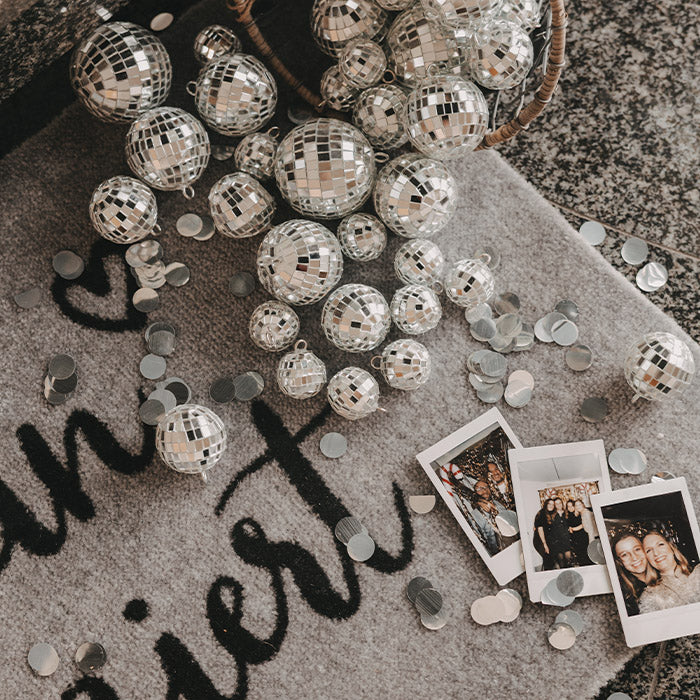 Graue Fußmatte mit schwarzem Schriftzug „Hereinspaziert“, dekoriert mit silbernen Discokugeln, Konfetti und Polaroid-Fotos.