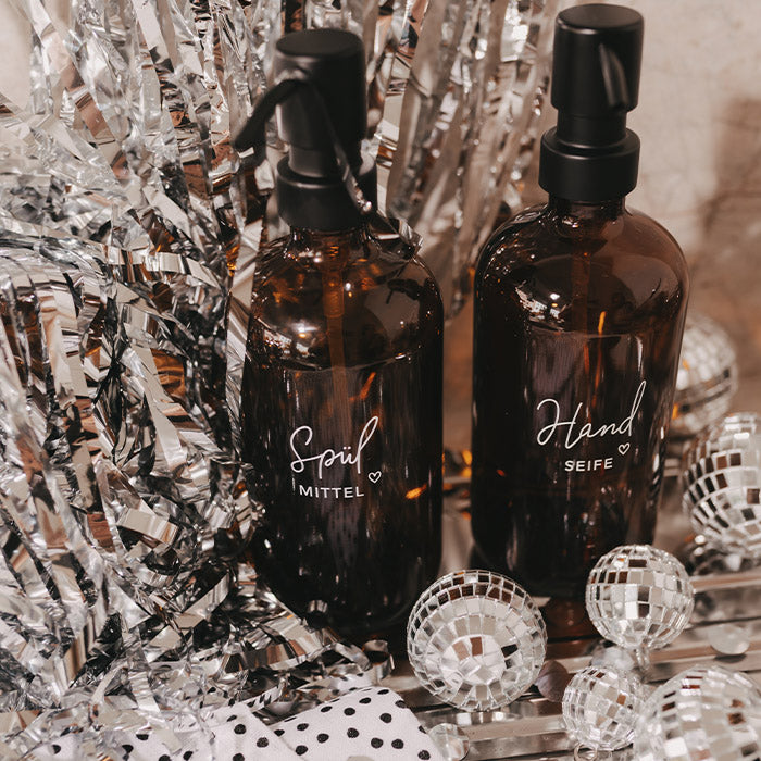 Zwei braune Glasflaschen mit schwarzen Pumpspendern und den Aufschriften „Spülmittel“ und „Handseife“, dekoriert mit silbernen Discokugeln und Lametta.