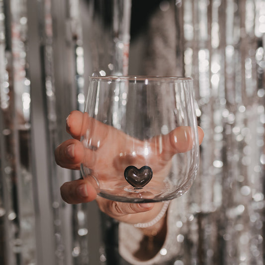 Transparentes Trinkglas mit kleiner schwarzer Herzfigur im Inneren, von einer Hand vor einem glitzernden silbernen Partyhintergrund gehalten.