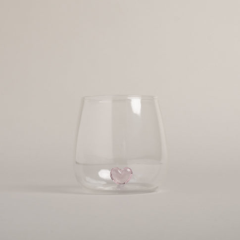 Klares Trinkglas mit rosa Herzfigur am Boden, modernes Design auf hellem Hintergrund fotografiert.