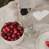Glas-Trinkflasche mit schwarzem Herz neben Schale mit Kirschen und Kühltasche beim Picknick