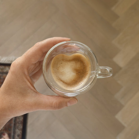 ine Hand hält eine transparente Glas-Tasse mit Cappuccino. Die Crema auf dem Kaffee hat eine natürliche Herzform. Der Hintergrund zeigt einen hellen Holzboden in Fischgrätmuster. Die Szene vermittelt Wärme, Gemütlichkeit und Achtsamkeit – perfekt für einen liebevollen Start in den Tag.