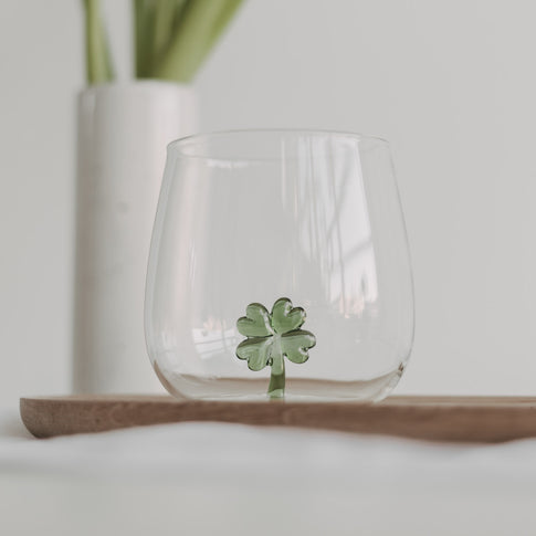 Trinkglas mit einer grünen Kleeblattfigur im Glasboden, auf einem Holztablett platziert, heller Hintergrund.
