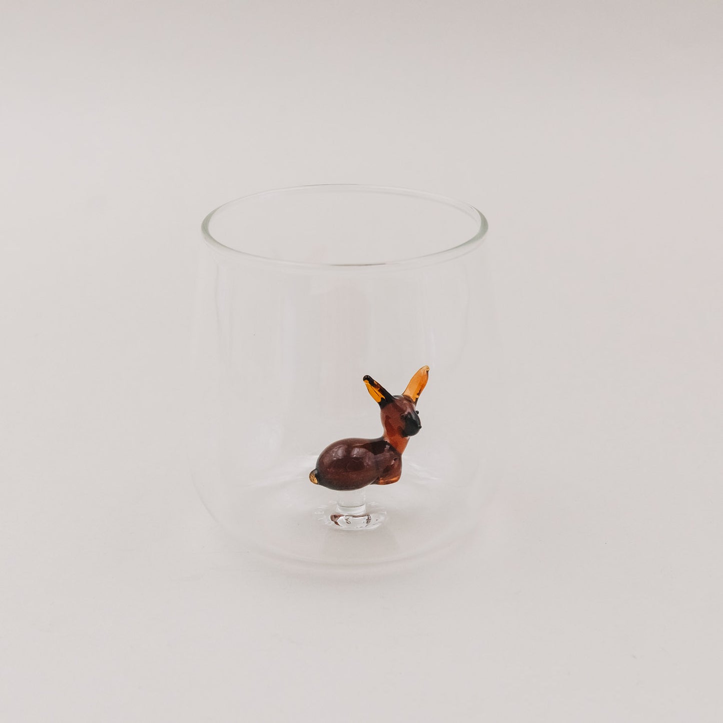 Trinkglas mit Rehfigur aus braunem Glas auf hellem Hintergrund – das Reh steht zentriert im Glas.