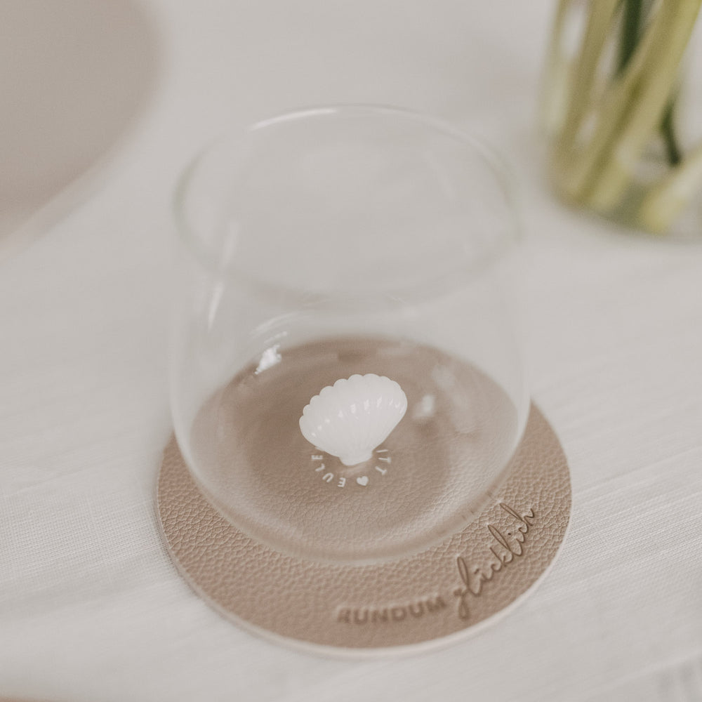 Trinkglas mit Muschelmotiv am Boden auf beigem Untersetzer mit Prägung