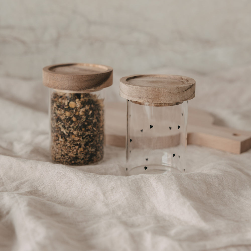 Zwei Vorratsgläser mit Holzdeckel, eines mit Gewürzen gefüllt, eines mit schwarzen Herzen