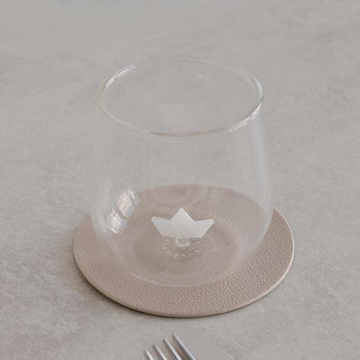Transparentes Glas mit kleinem Papierboot im Inneren, dekorativ auf hellem Untergrund mit Geschirr arrangiert