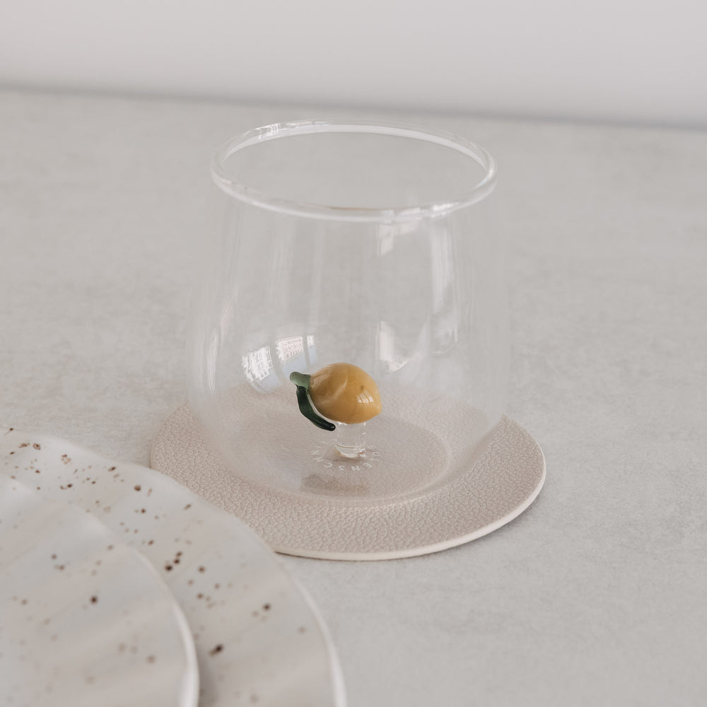 Transparentes Trinkglas mit kleiner Zitrone im Inneren, stilvoll inszeniert auf hellem Untergrund neben Geschirr