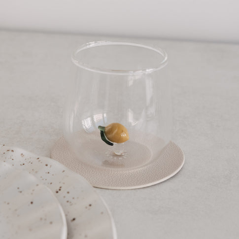 Transparentes Trinkglas mit kleiner Zitrone im Inneren, stilvoll inszeniert auf hellem Untergrund neben Geschirr