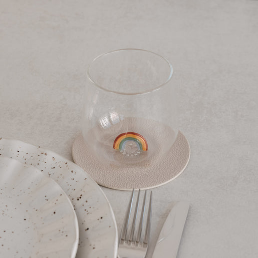 Trinkglas mit Regenbogen-Motiv innen, auf Untersetzer neben Teller und Besteck, minimalistisches Setting