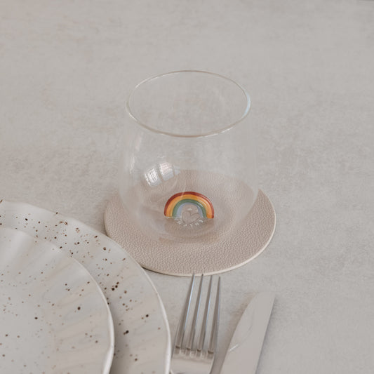 Trinkglas mit Regenbogen-Motiv innen, auf Untersetzer neben Teller und Besteck, minimalistisches Setting