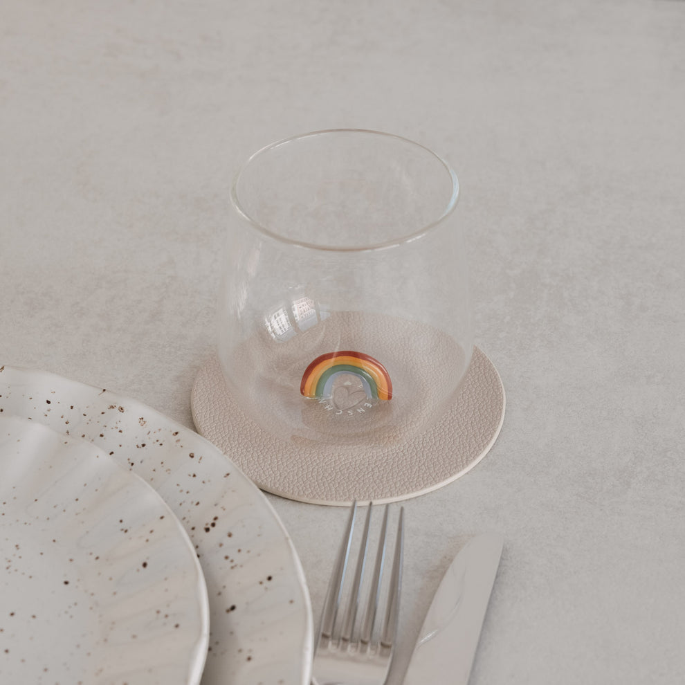 Trinkglas mit Regenbogen-Motiv innen, auf Untersetzer neben Teller und Besteck, minimalistisches Setting
