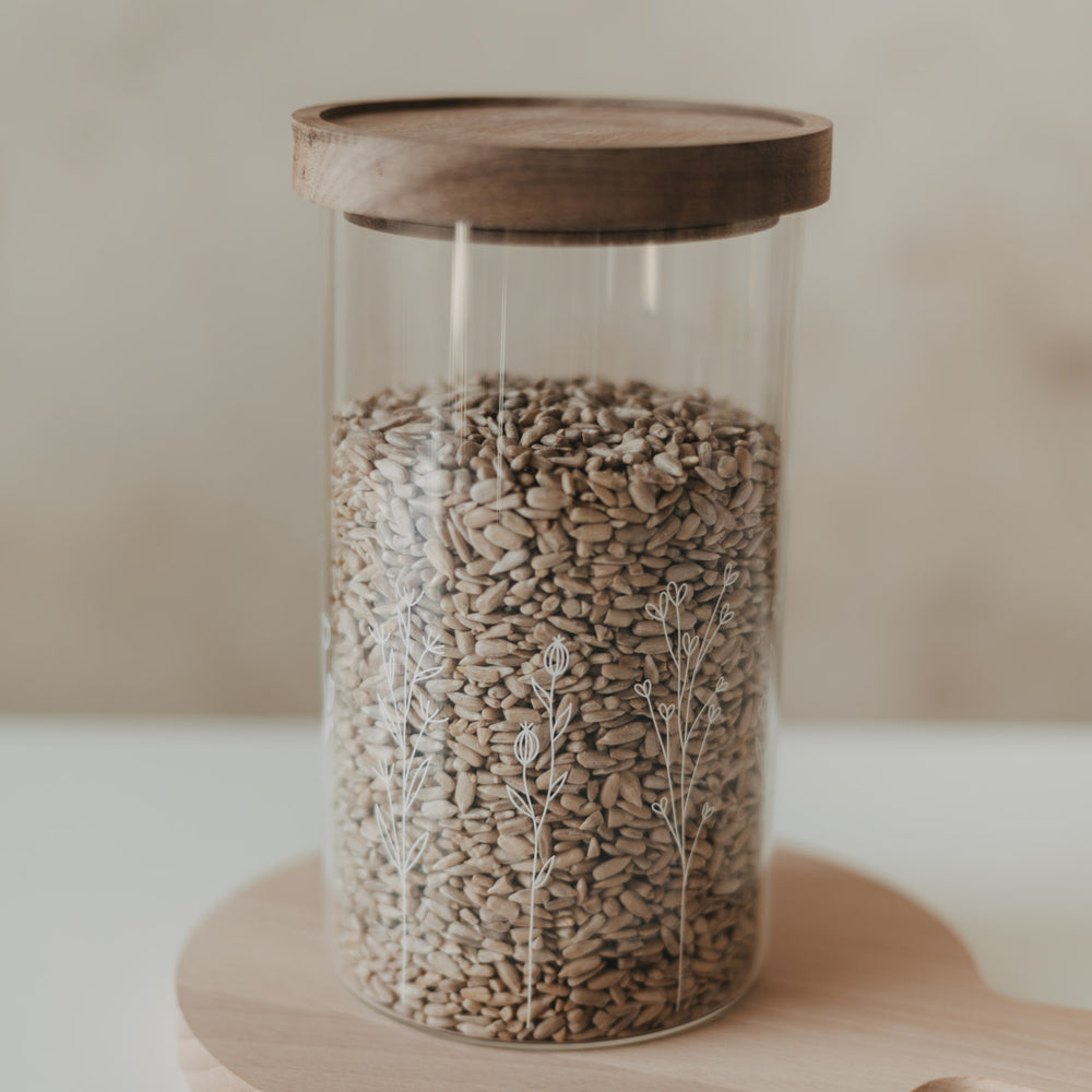 Glas-Vorratsbehälter mit feinem weißem Blumenmotiv und dunklem Holzdeckel, gefüllt mit Sonnenblumenkernen
