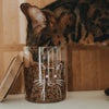 Nahaufnahme einer getigerten Katze beim Fressen aus einem Vorratsglas mit weißem Katzenmotiv.