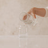 Hand gießt Wasser aus Glas Karaffe in kleine Vase mit Herzmotiv