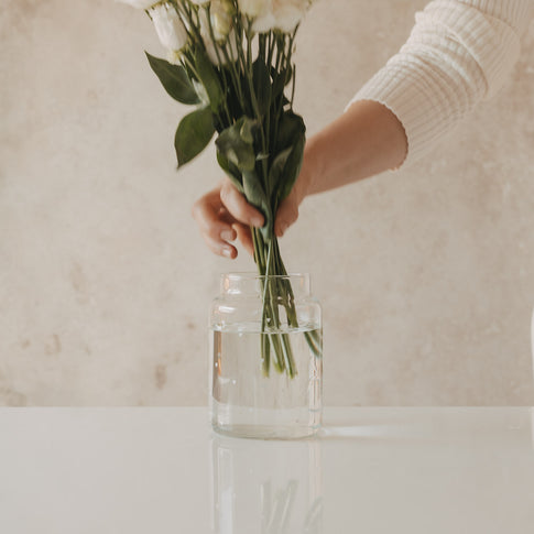 Hand stellt weißen Blumenstrauß in Glasvase mit Herzmotiv