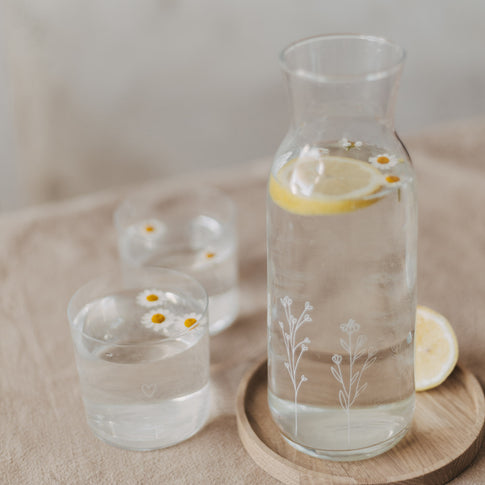 Glas Karaffe mit Zitronenwasser und zwei Gläsern mit Blüten auf Holztablett