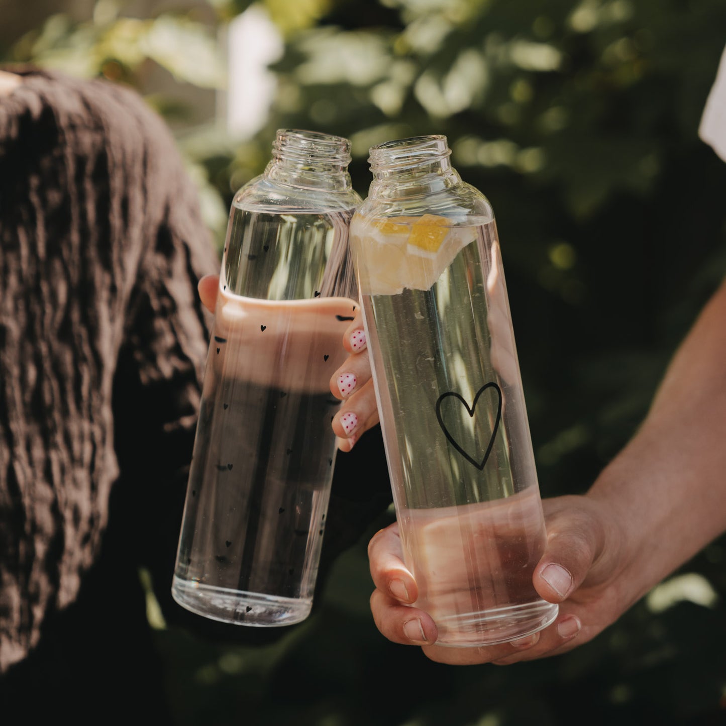 Zwei Trinkflaschen aus Glas mit Herzmotiv und Zitronenwasser in der Hand
