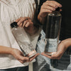 Zwei Glas Trinkflaschen im Vergleich, eine mit Herzmotiv, eine mit floralem Muster, in Händen gehalten