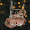 Transparentes Trinkglas mit rot-weißer Zuckerstange im Glasboden, gefüllt mit Wasser vor warmen Weihnachtslichtern.
