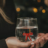 Transparentes Wasserglas mit roter Schleife im Glasboden, gehalten vor stimmungsvollen Weihnachtslichtern.