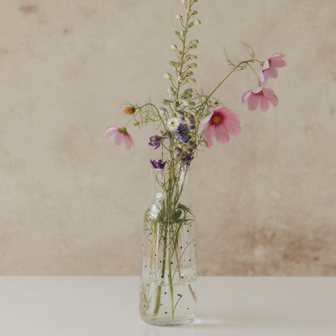 Glaskaraffe mit kleinen schwarzen Herzen als Vase mit rosa und violetten Blumen