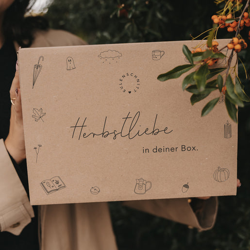 Nahaufnahme der Herbstbox mit Illustrationen und dem Text „Herbstliebe in deiner Box“ vor Beerenzweigen.