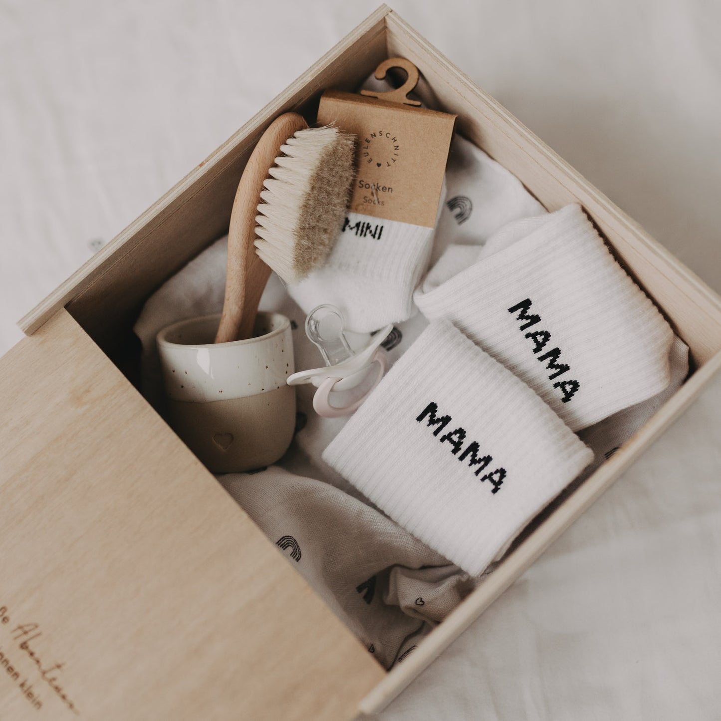 Geöffnete Erinnerungsbox aus Holz, gefüllt mit Babyartikeln wie Mini-Söckchen, Bürste und Schnuller.