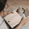 Person hält eine geöffnete Erinnerungsbox aus Holz in den Händen. Im Inneren befinden sich ein Keramikbecher, Trockenblumen, eine Karte und weiche Socken mit Herzmuster – liebevoll arrangiert als Geschenkset.