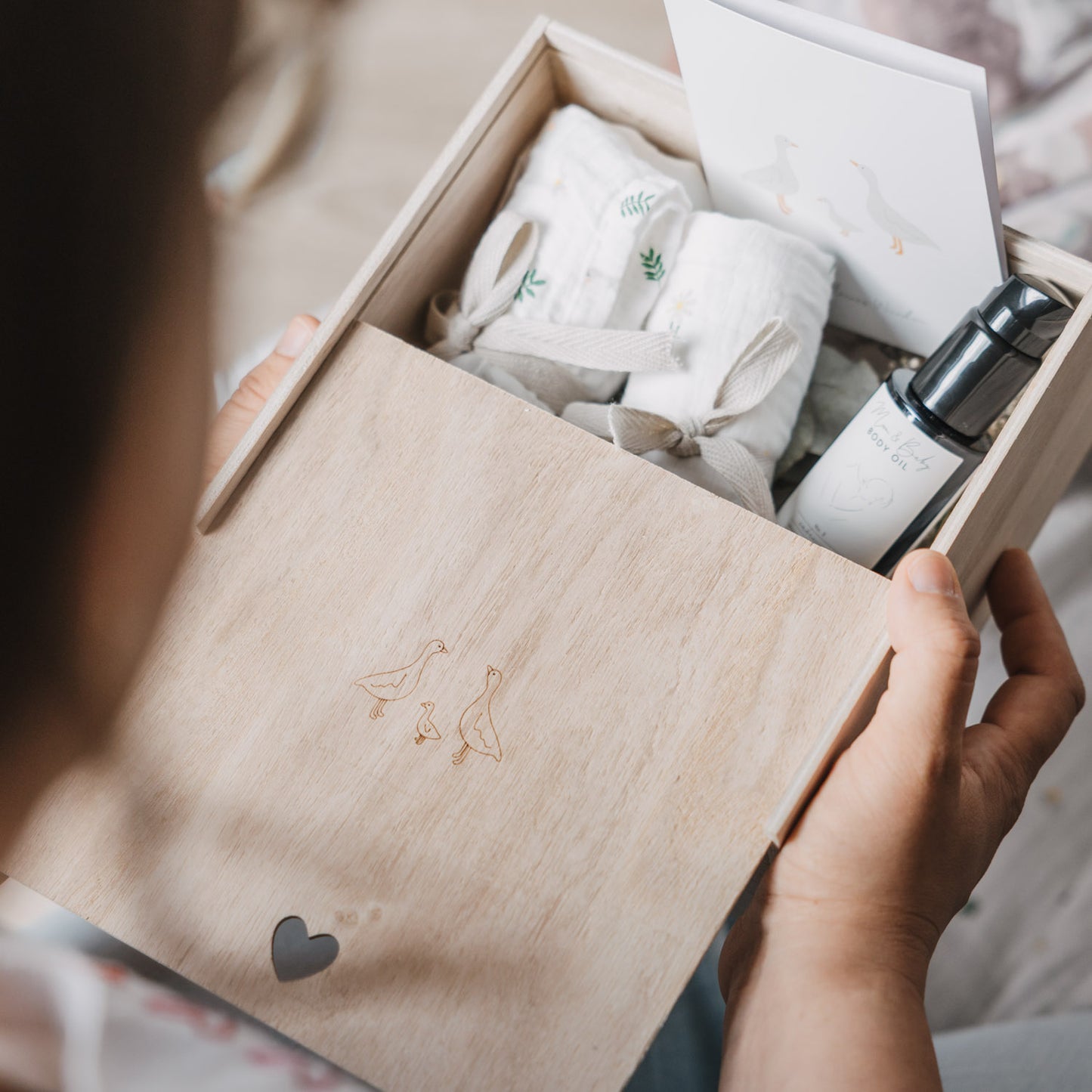 Person hält eine geöffnete Erinnerungsbox aus hellem Holz mit eingravierter Gänsefamilie in den Händen. Im Inneren befinden sich liebevoll arrangierte Musselintücher, eine Grußkarte mit Gänsemotiv und ein Pflegeöl für Mama und Baby.