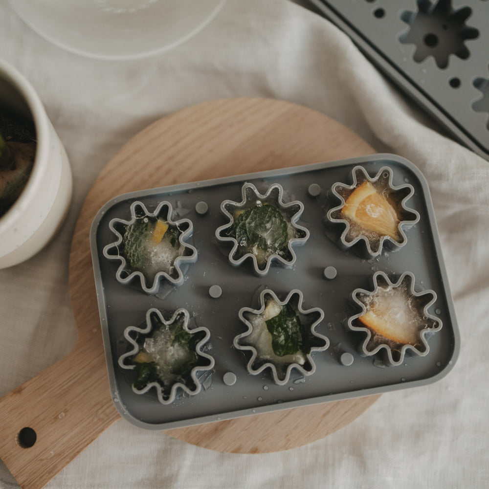 Fertige Eiswürfel mit Orangenscheiben oder Minze liegen in der grauen Blütenform aus flexiblem Silikon, dekoriert auf einem runden Holzbrett.