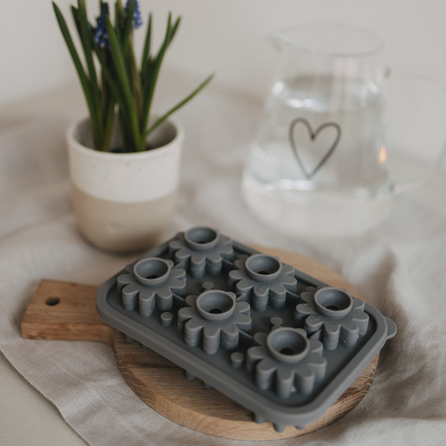 Grauen Blütenform aus flexiblem Silikon liegt dekoriert auf einem runden Holzbrett, dahinter ein Becher gefüllt mit einer Pflanze und eine Karaffe gefüllt mit Wasser.