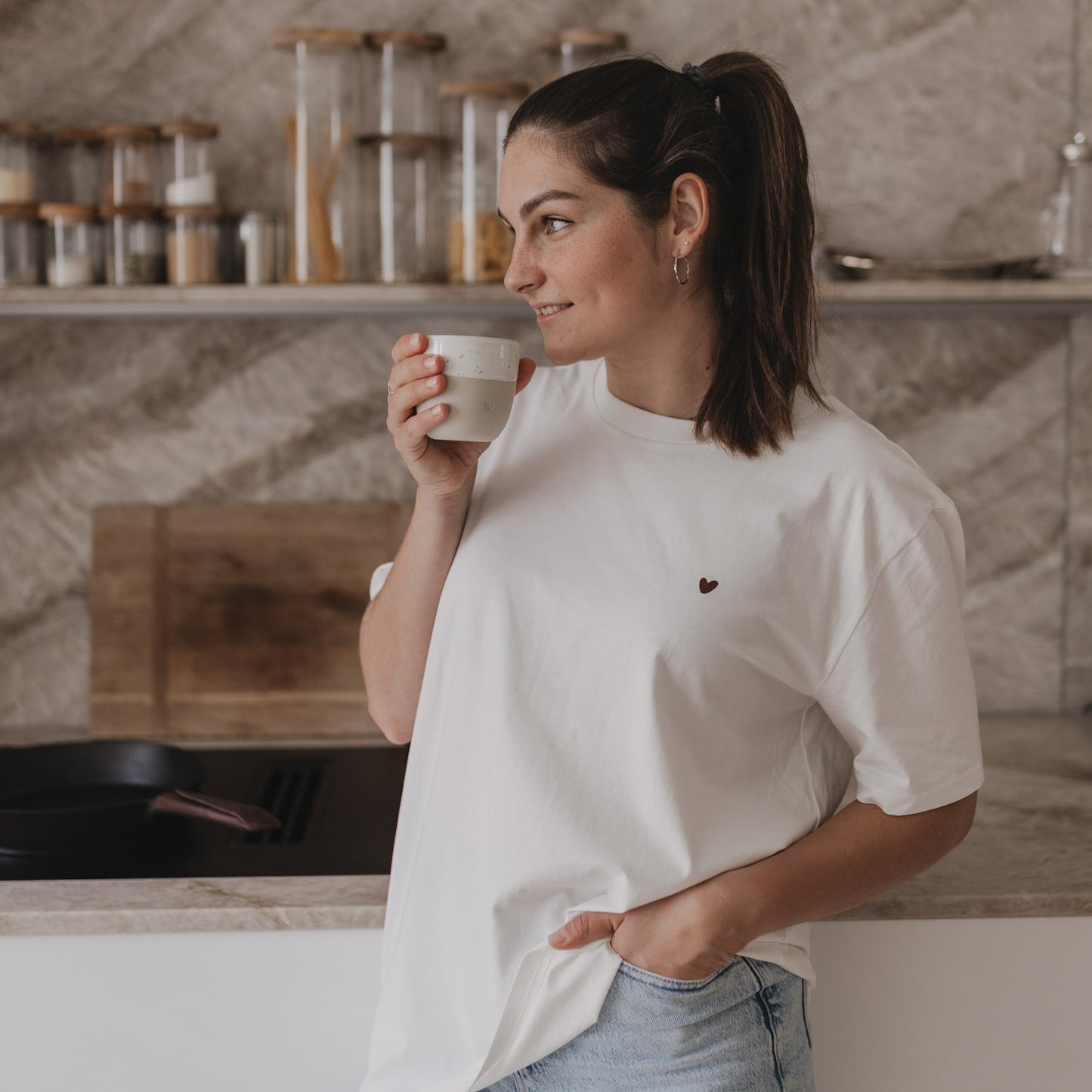 Frau lächelt mit Kaffeetasse in der Hand, trägt weißes Shirt mit Herzprint auf der Brust.