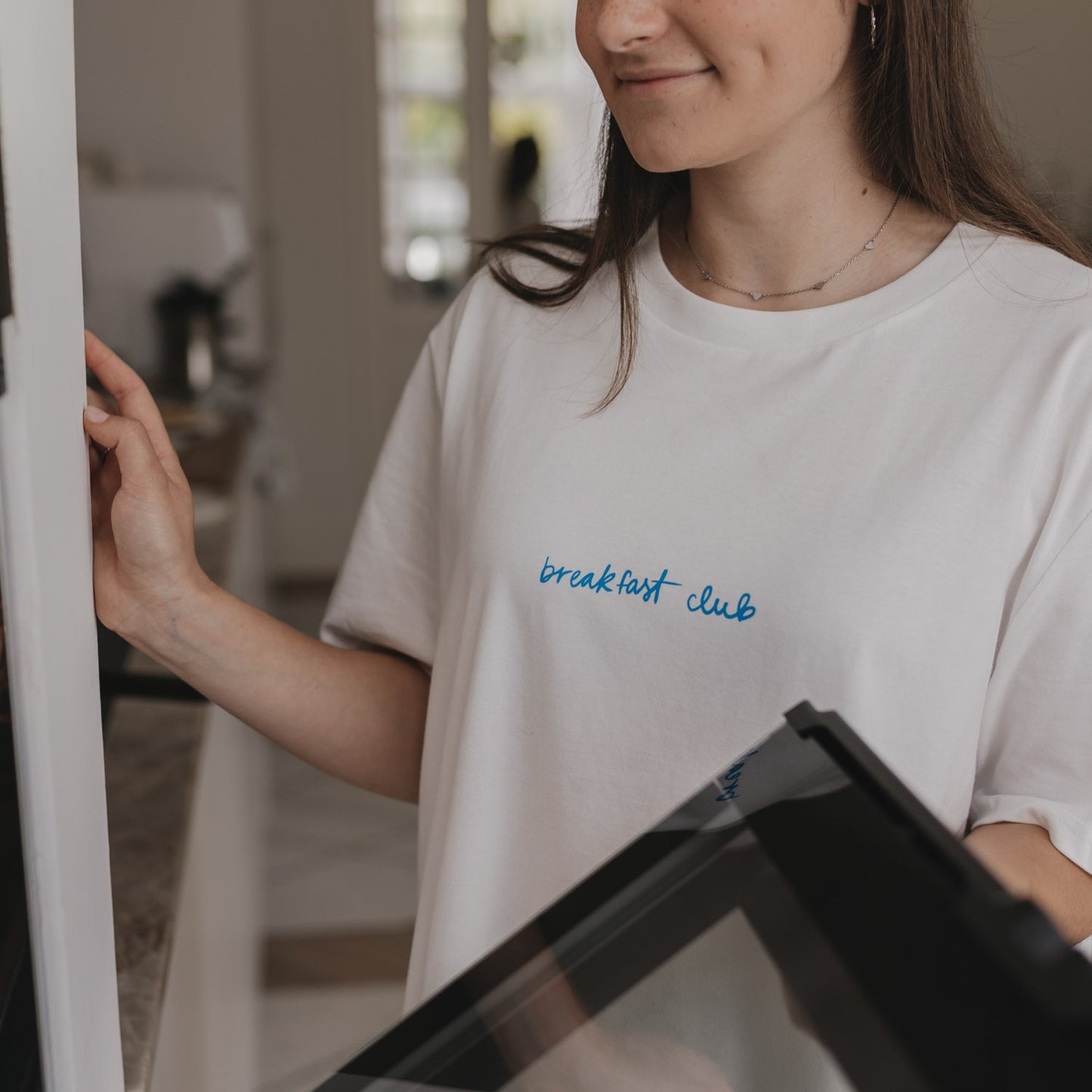 Frau öffnet Ofentür, trägt weißes Shirt mit blauem „breakfast club“-Schriftzug.