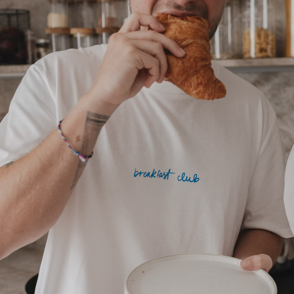 Mann im weißen Breakfast Club Shirt beißt in Croissant – Fokus auf blauen Frontprint.
