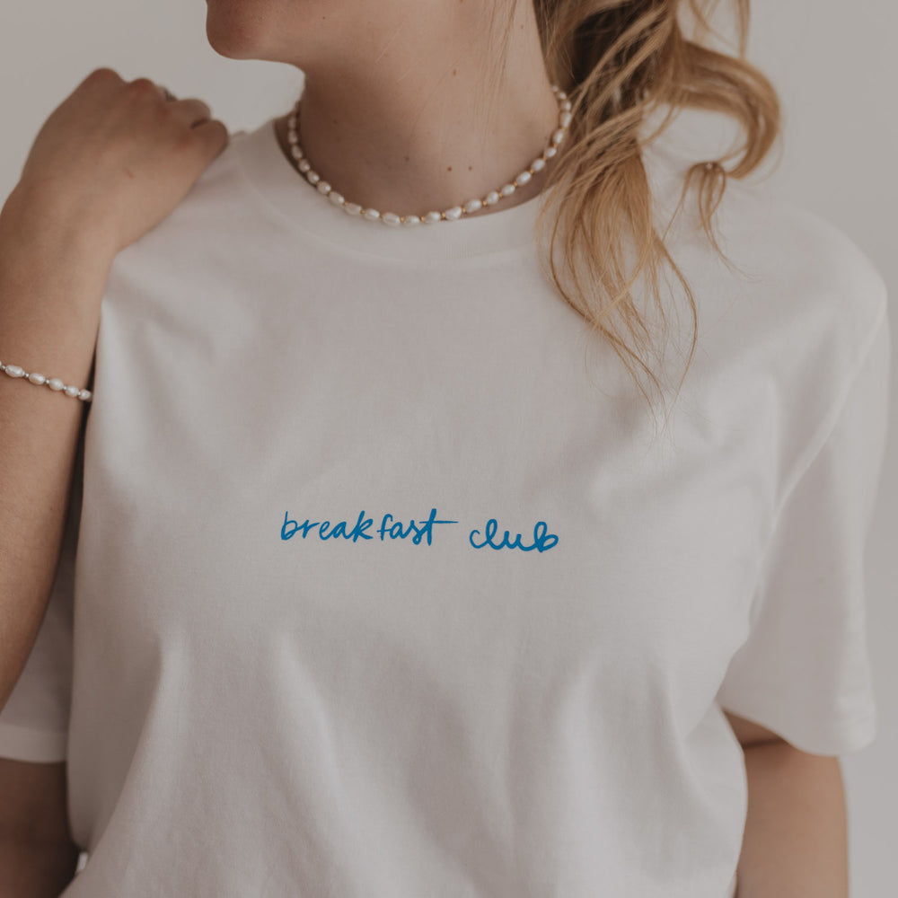 Nahaufnahme des blauen „breakfast club“ Prints auf weißem T-Shirt, getragen von Frau mit Perlenkette.