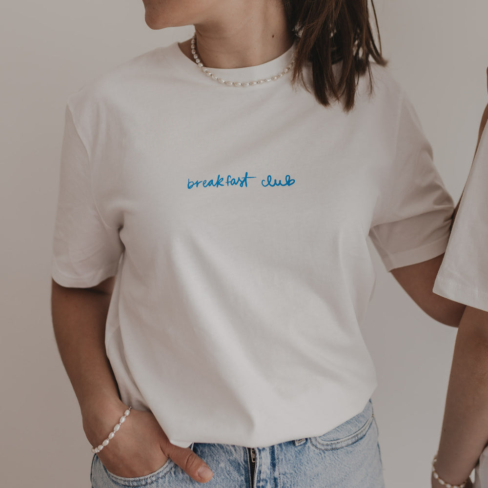Frau trägt weißes T-Shirt mit „breakfast club“-Print in Blau – moderner Look mit Perlenkette und Jeans.