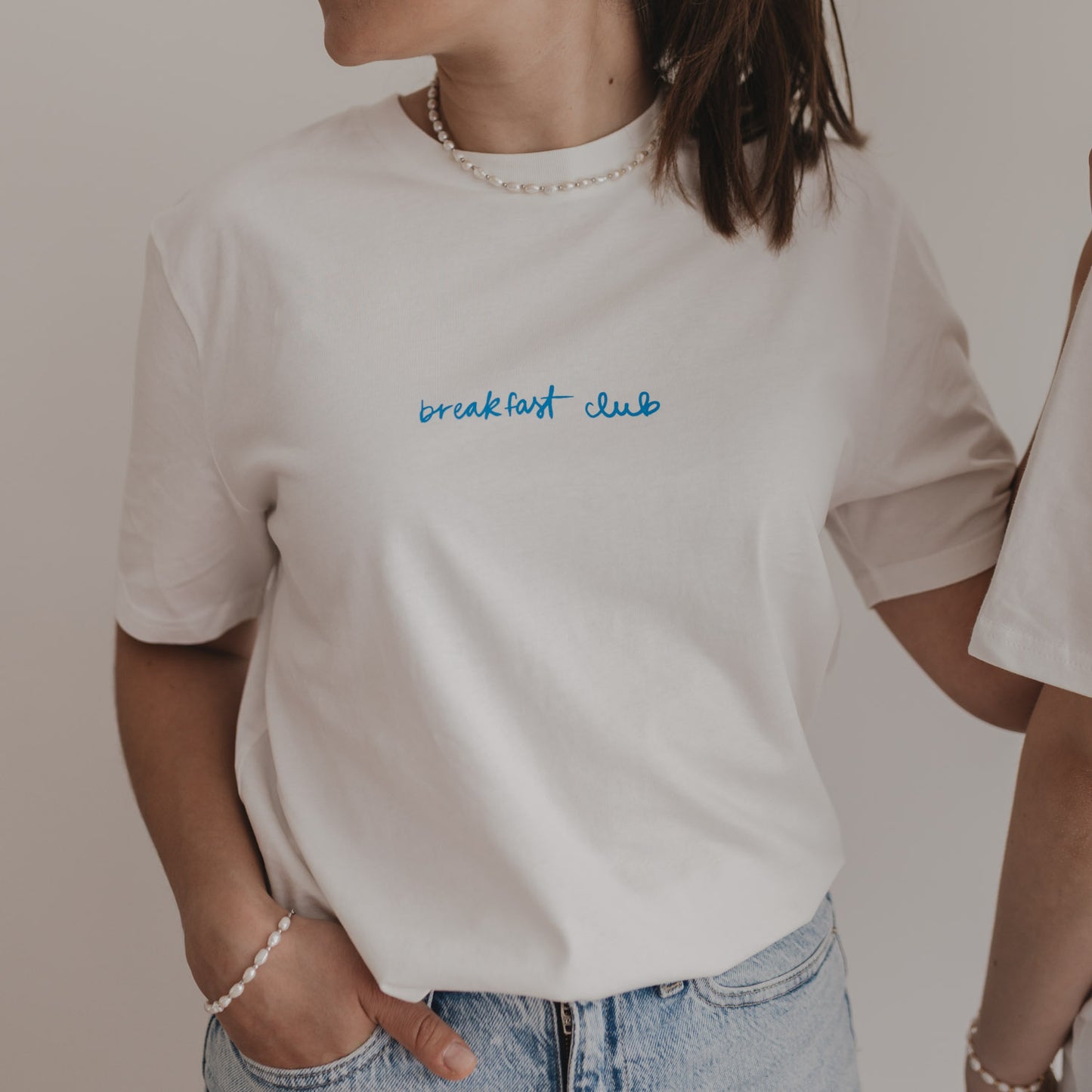 Frau trägt weißes T-Shirt mit „breakfast club“-Print in Blau – moderner Look mit Perlenkette und Jeans.