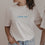 Frau trägt weißes T-Shirt mit „breakfast club“-Print in Blau – moderner Look mit Perlenkette und Jeans.